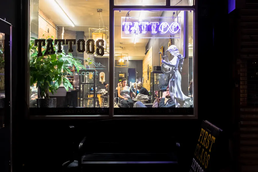The shop window at night, lit up, with a neon tattoo sign.